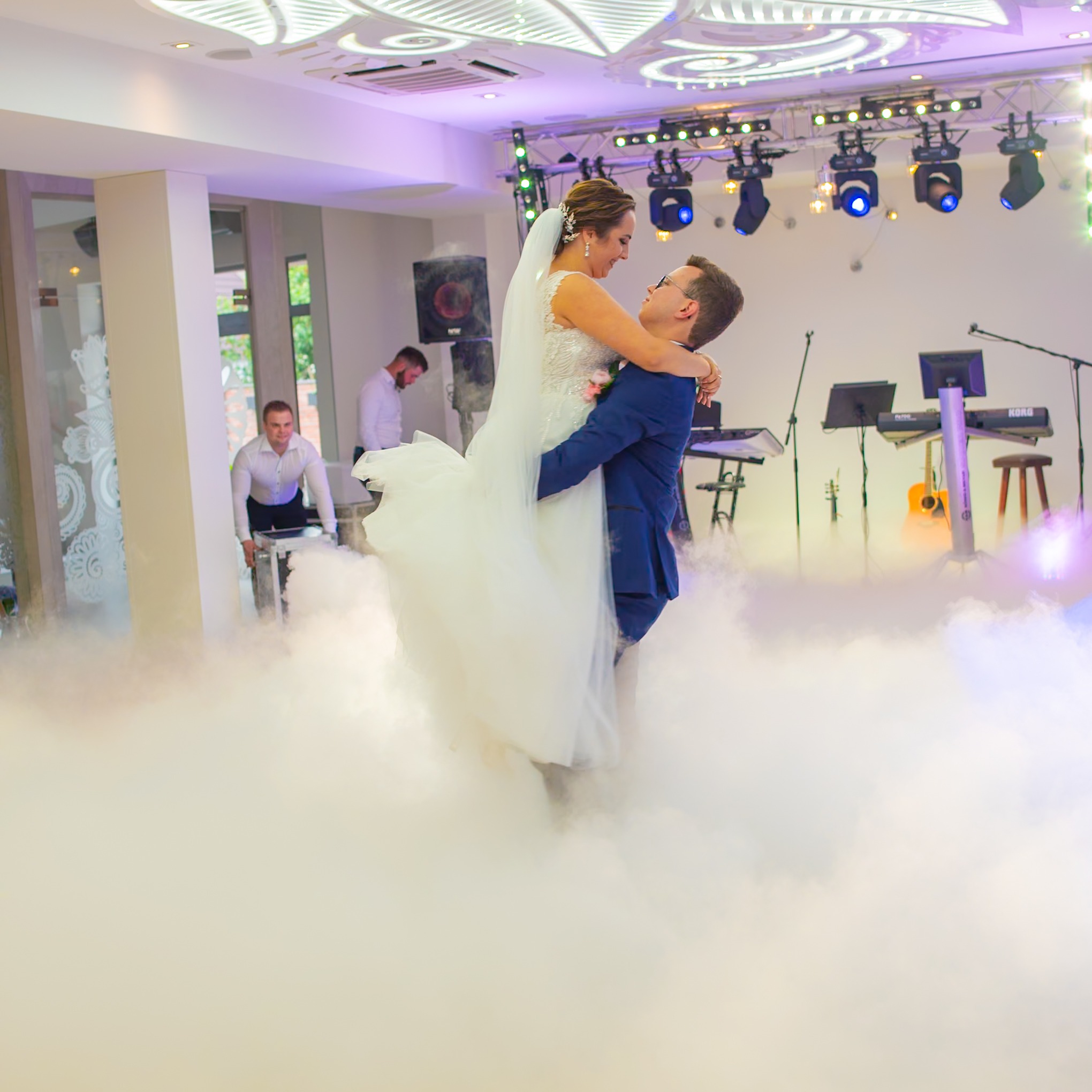 Uśmiechnięta para młoda w objęciach na parkiecie podczas pierwszego tańca, otoczona sztuczną mgłą, w tle oświetlenie sceniczne i zespół muzyczny.