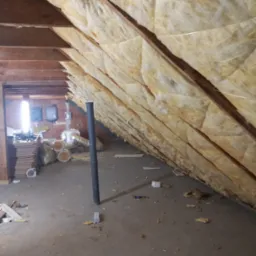 Poddasze w trakcie ocieplania wełną mineralną, widoczne drewniane krokwie i fragment instalacji wentylacyjnej, na podłodze resztki materiałów i narzędzi.