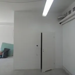 Wnętrze w trakcie remontu: gładkie, białe ściany, otwarte białe drzwi, widoczne instalacje elektryczne, materiały budowlane i narzędzia w tle, surowy beton na podłodze, oświetlenie sufitowe.