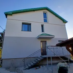 Dwukondygnacyjny dom z ocieploną elewacją, świeżo pomalowany na biało i szaro, z zielonym dachem i schodami wejściowymi, z widocznym rusztowaniem po lewej stronie i psem rasy labrador w ogrodzie.