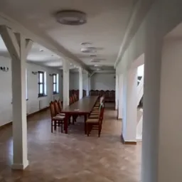 Jasne, przestronne wnętrze sali z drewnianymi stołami i krzesłami, po remoncie. Białe ściany i filary z drewnianymi elementami. Widoczne oświetlenie sufitowe i okna.