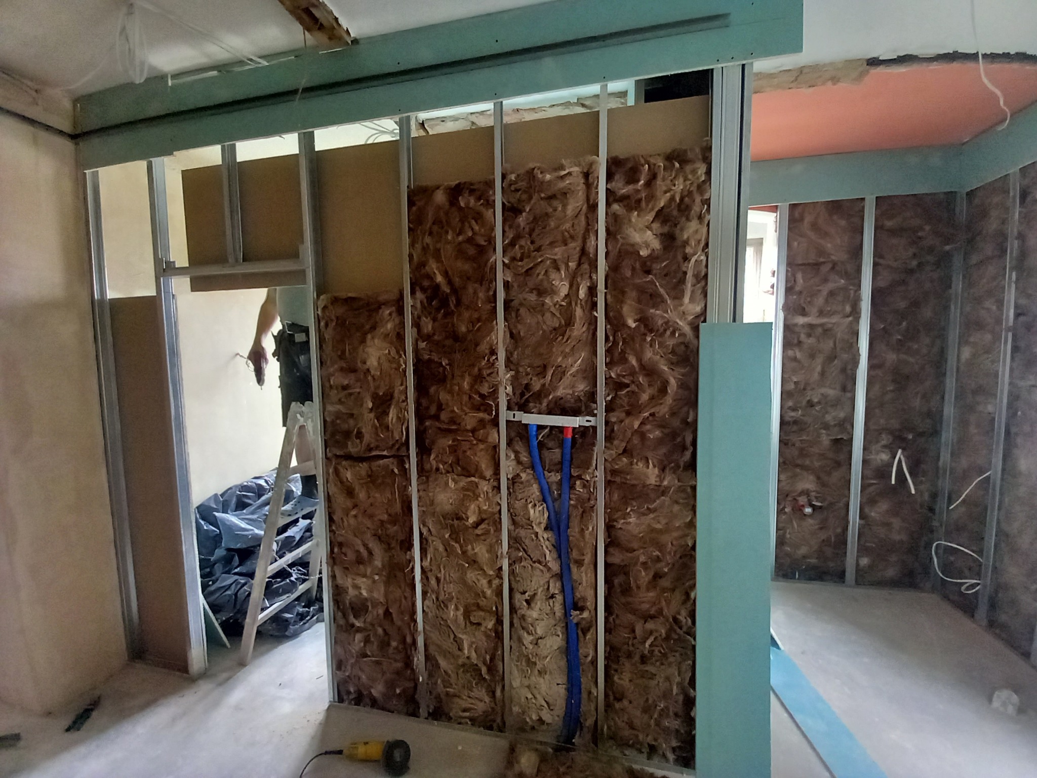 Wnętrze w trakcie prac: montaż metalowej konstrukcji, wypełnienie wełną mineralną, widoczne niebieskie rury instalacyjne, drabina i narzędzia na podłodze.