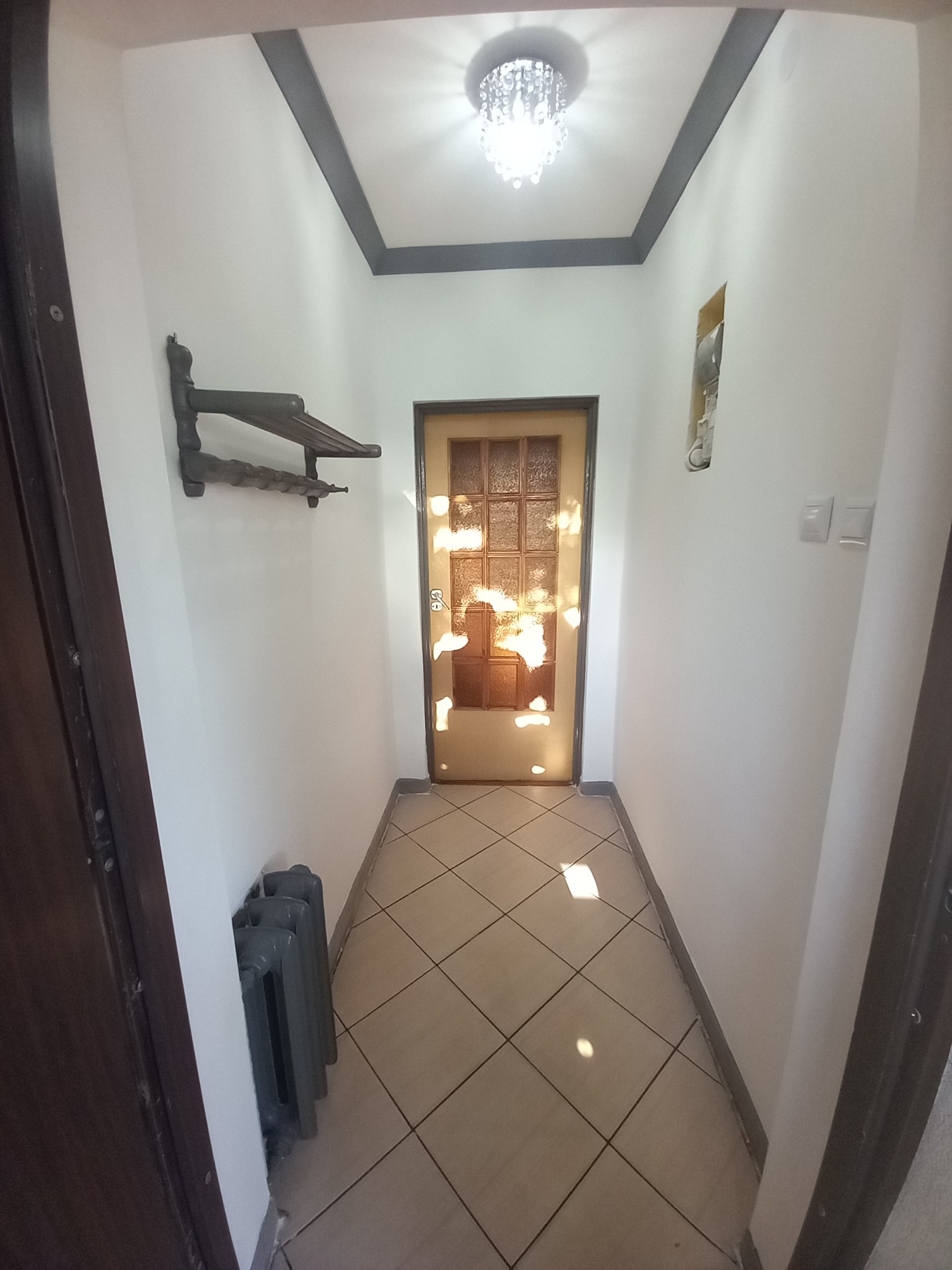 Wnętrze korytarza po remoncie: gładkie, białe ściany z szarą lamperią, brązowa podłoga z płytek, drewniany wieszak na ubrania, grzejnik żeliwny, drzwi z szybą ornamentową i kryształowy żyrandol.