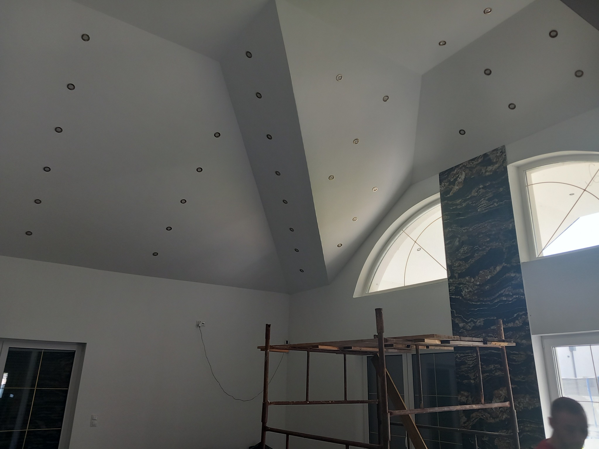Wnętrze domu w trakcie wykończenia: białe ściany i sufit z zamontowanymi punktowymi lampami, drewniane rusztowanie, okno łukowe i dekoracyjny panel ścienny z ciemnego kamienia.