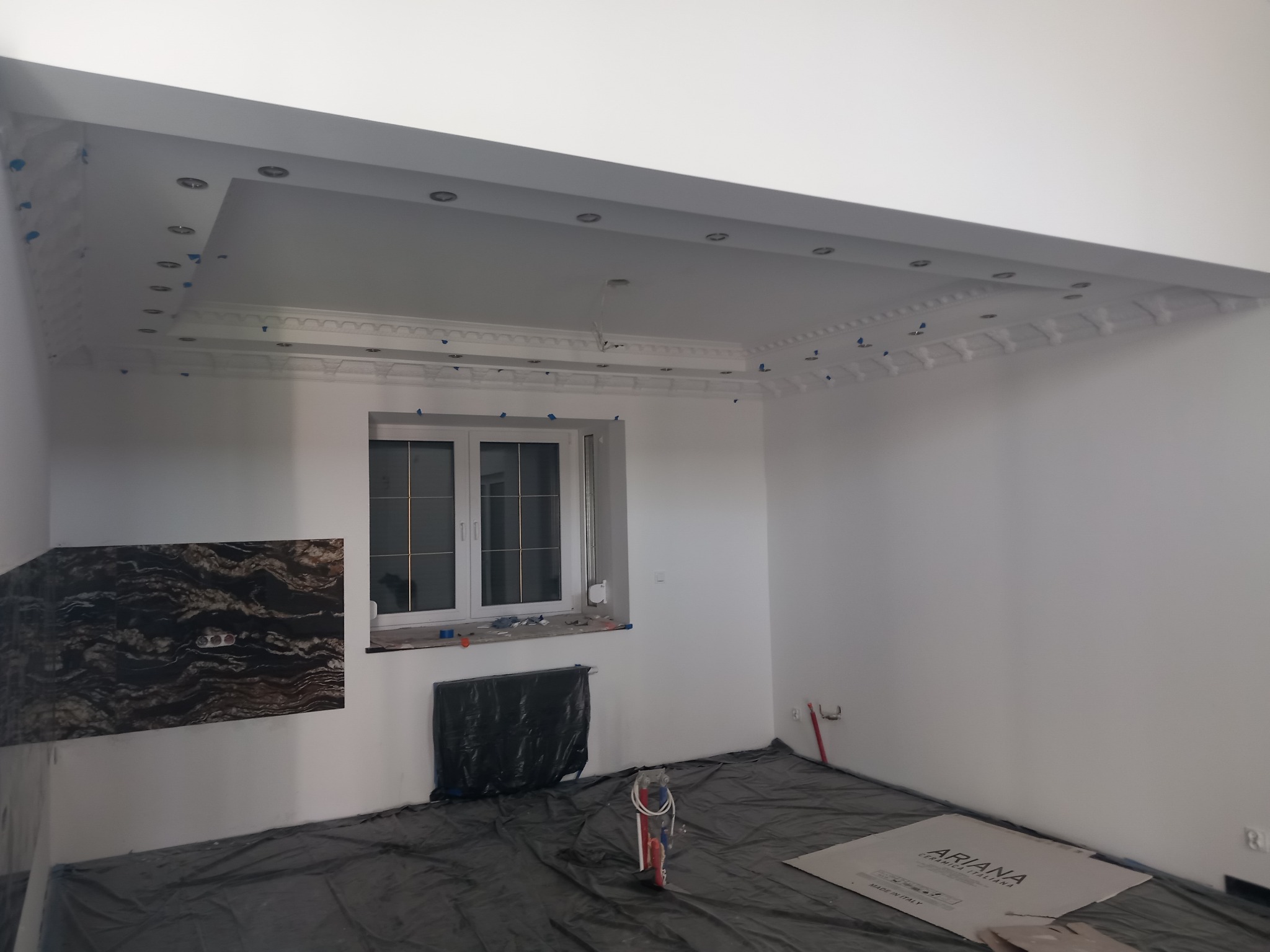 Pokój w trakcie remontu: sufit z dekoracyjnymi listwami i punktowym oświetleniem, zabezpieczona folią podłoga, okno z grzejnikiem pod spodem, fragment ściany z okładziną imitującą marmur.
