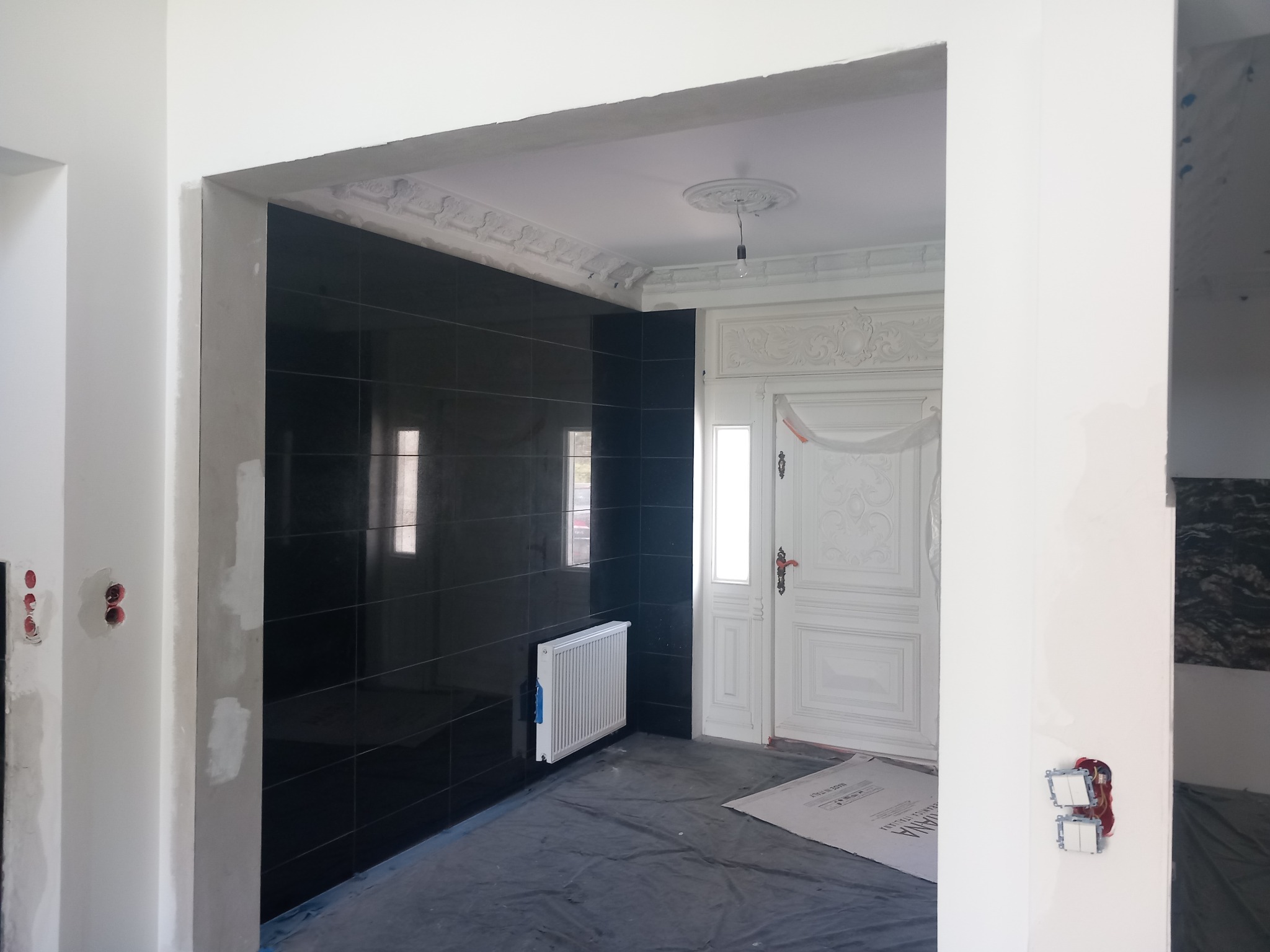 Wnętrze w trakcie prac wykończeniowych: czarne płytki na ścianie, białe ściany w trakcie szpachlowania, dekoracyjny sufit z rozetą i listwami, zabezpieczona folią podłoga, grzejnik na ścianie.
