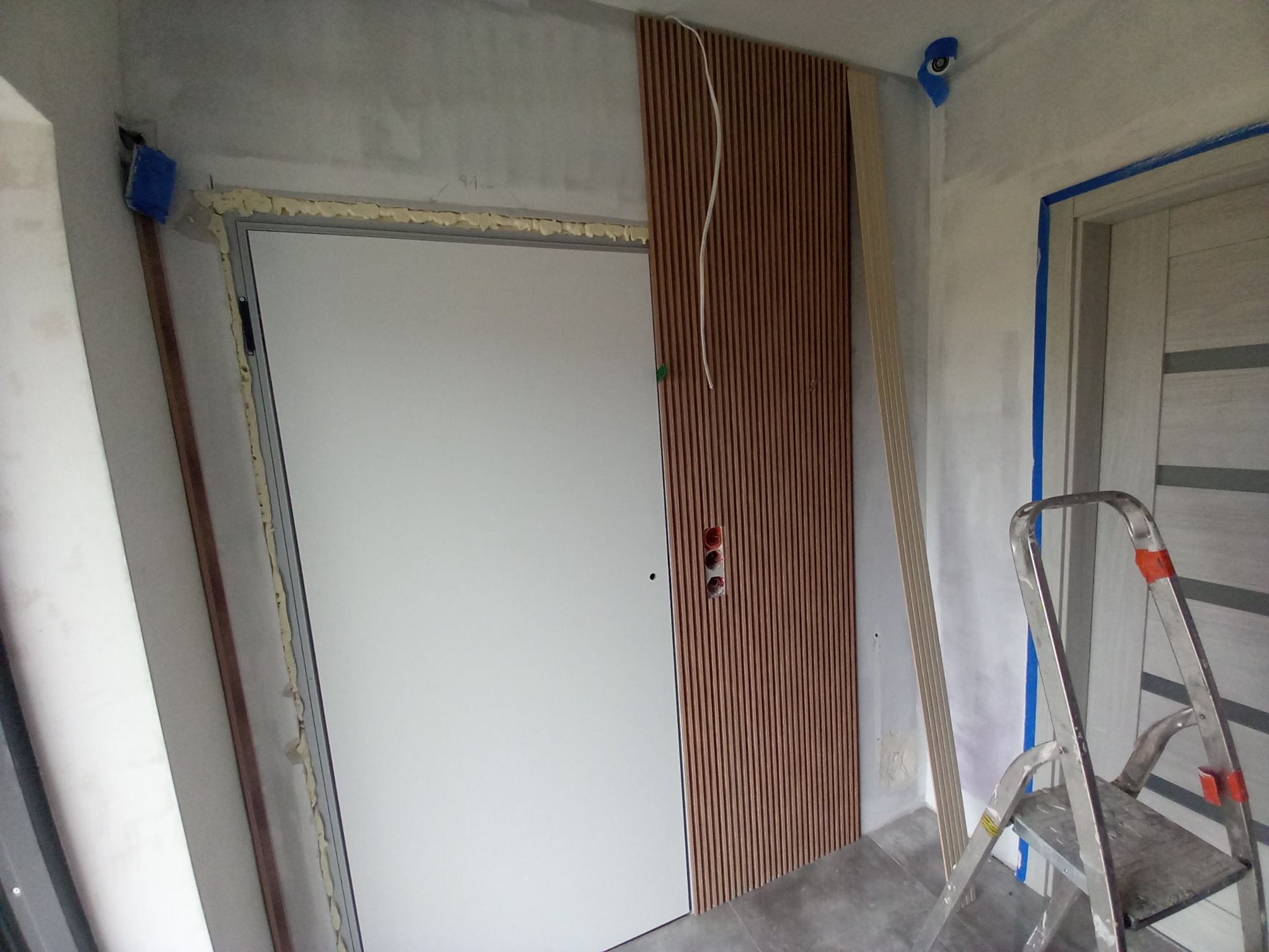 Montaż paneli ściennych z lameli drewnianych obok ościeżnicy drzwiowej zabezpieczonej pianką montażową, widoczne przewody elektryczne i drabina aluminiowa.