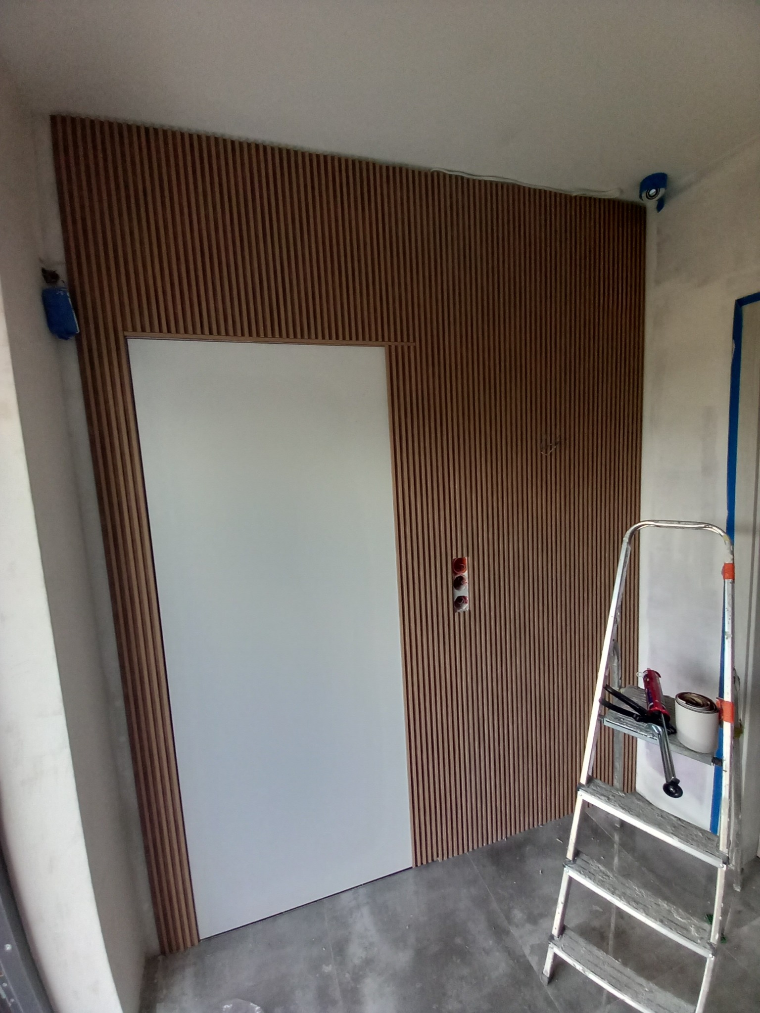 Montaż paneli ściennych z ukrytymi drzwiami, widoczna drabina i narzędzia, surowy stan wykończenia wnętrza.