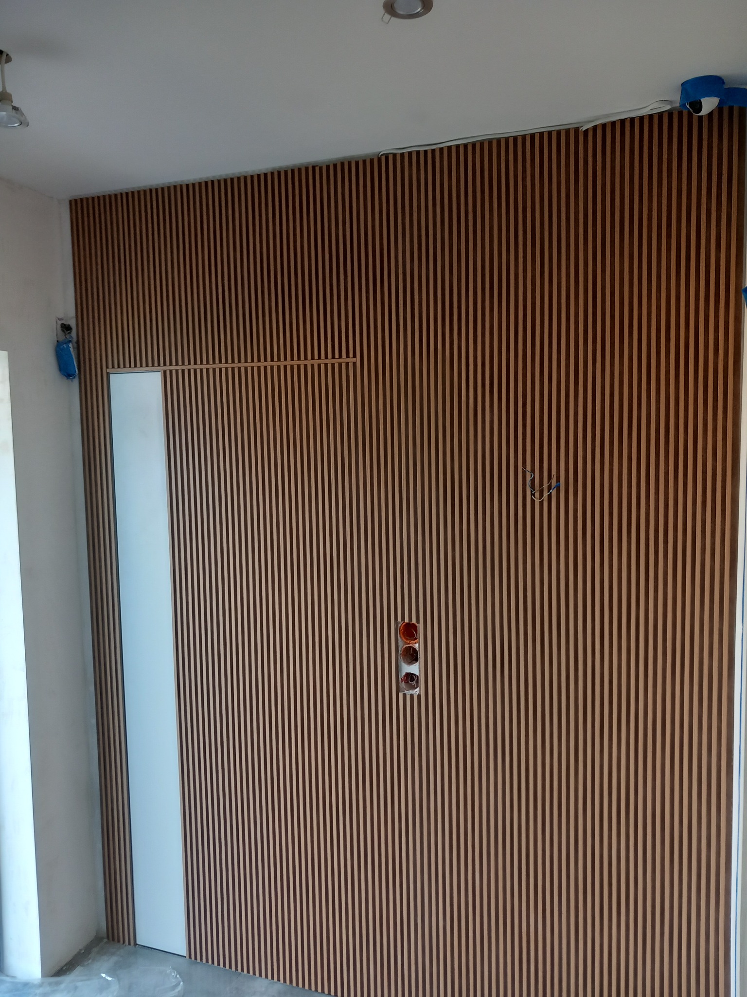 Drewniane lamele na ścianie z ukrytymi przewodami i gniazdkami, częściowo zakrywają białe drzwi.