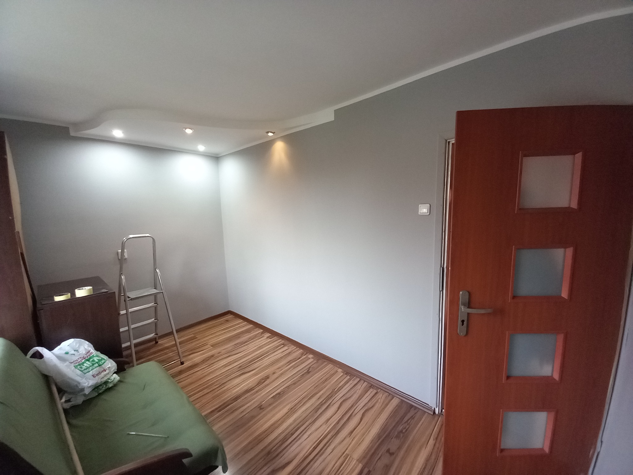 Pomieszczenie po remoncie: szare ściany, drewniana podłoga, częściowo widoczna drabina malarska i brązowe drzwi z trzema kwadratowymi przeszkleniami.