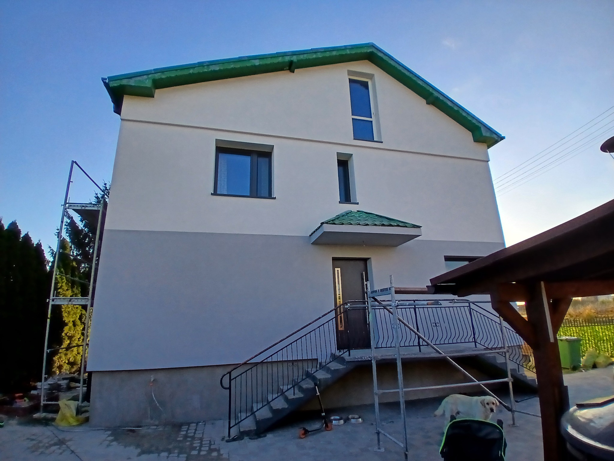 Dwukondygnacyjny dom z ocieploną elewacją, świeżo pomalowany na biało i szaro, z zielonym dachem i schodami wejściowymi, z widocznym rusztowaniem po lewej stronie i psem rasy labrador w ogrodzie.