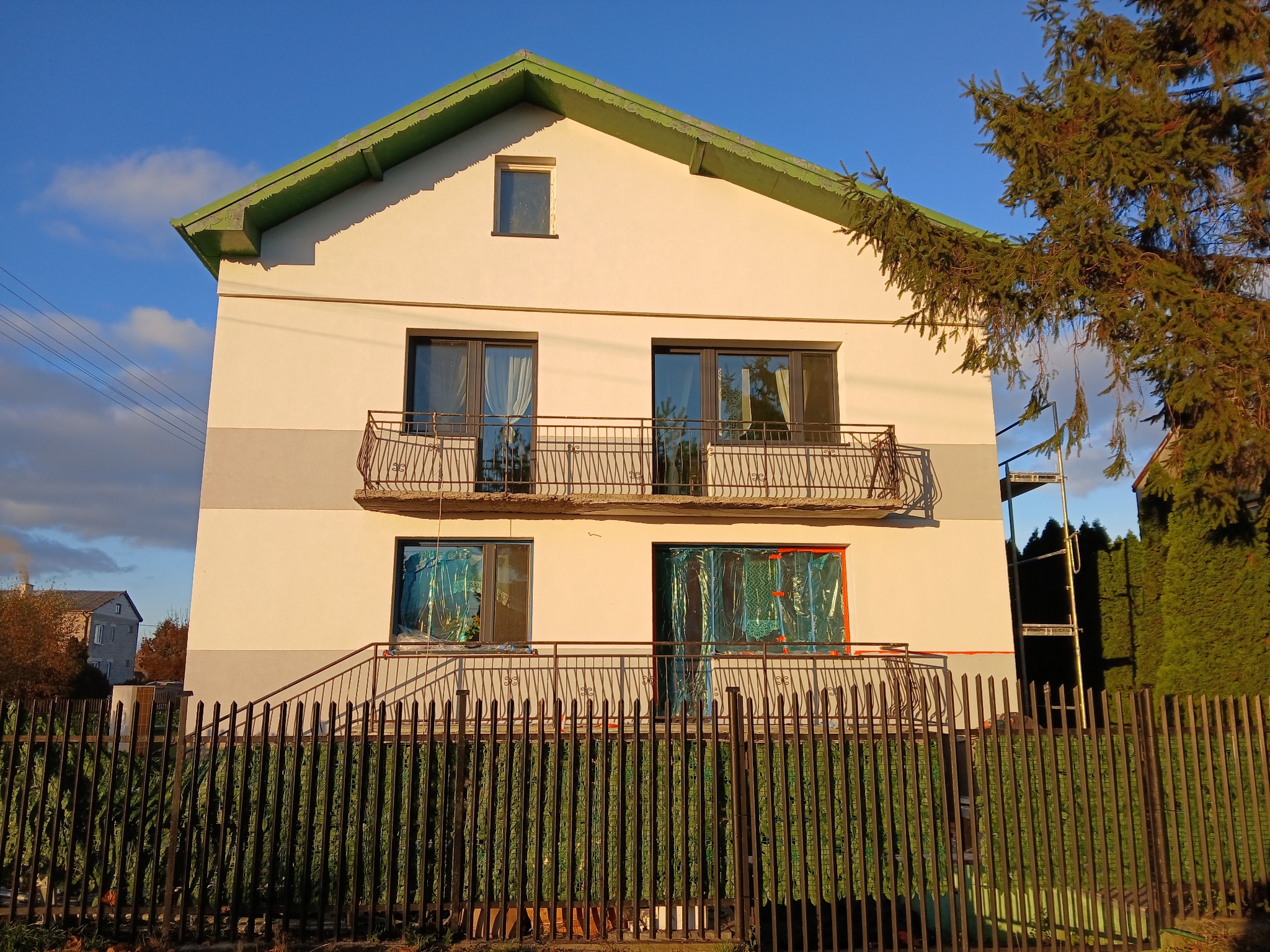 Dwupiętrowy dom z balkonami, okna zabezpieczone folią ochronną, sugerujące prace remontowe elewacji w trakcie realizacji, widoczne rusztowanie z boku budynku, metalowe ogrodzenie.