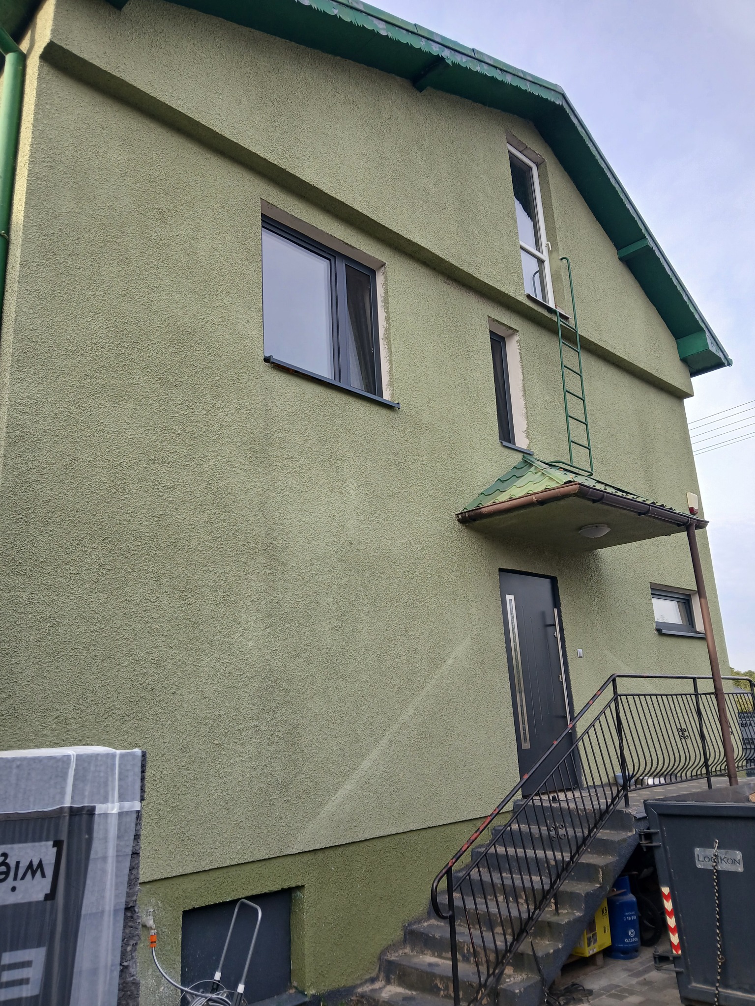 Zakończona renowacja elewacji domu z widoczną strukturą tynku baranka w odcieniach zieleni, ciemnoszare okna i drzwi, oraz metalowa drabina przymocowana do ściany.