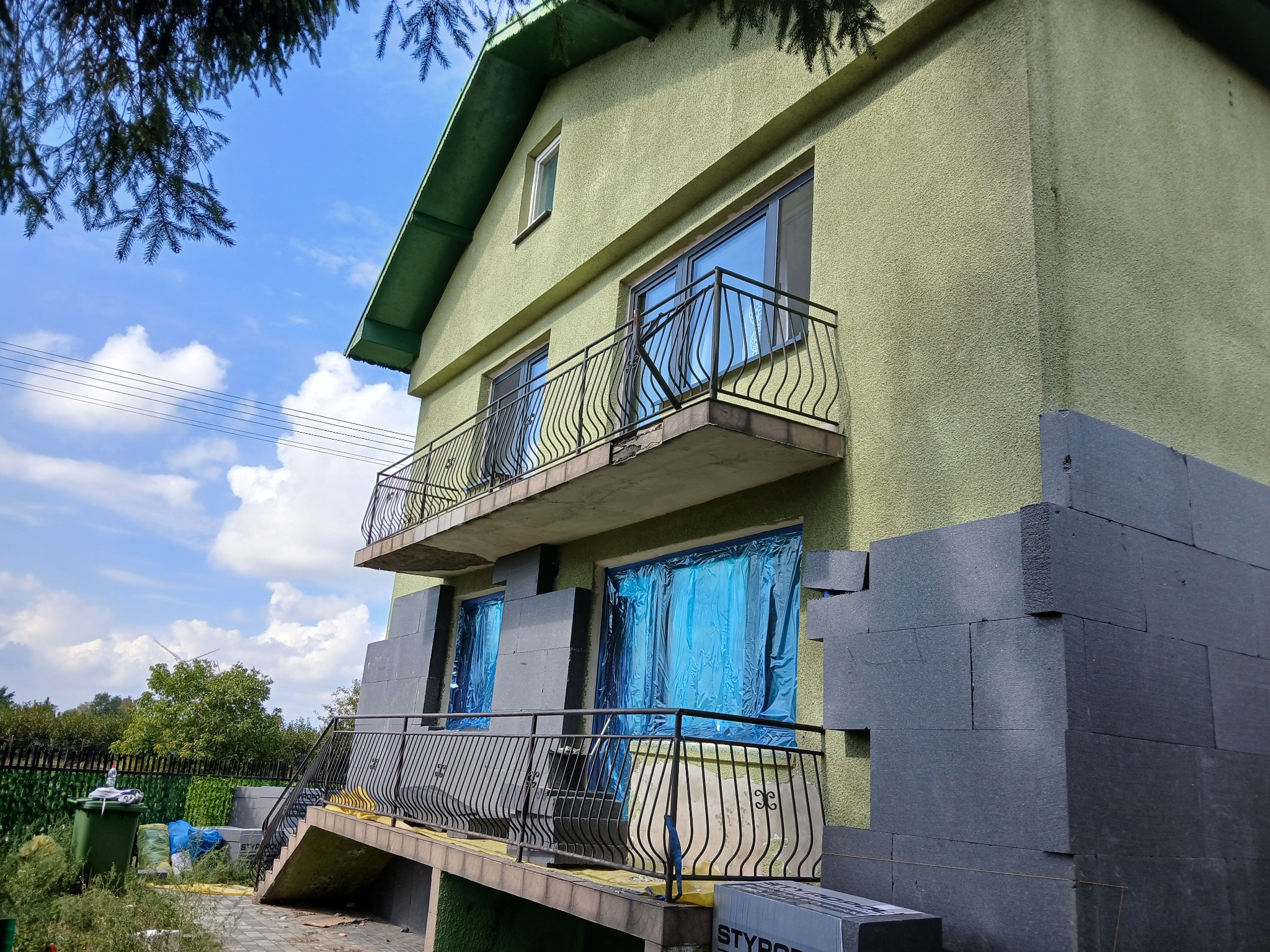 Dom jednorodzinny w trakcie nakładania izolacji termicznej elewacji, styropian grafitowy przyklejony do ściany, okna zabezpieczone folią, widok z dołu na balkon i fragment ogrodu.
