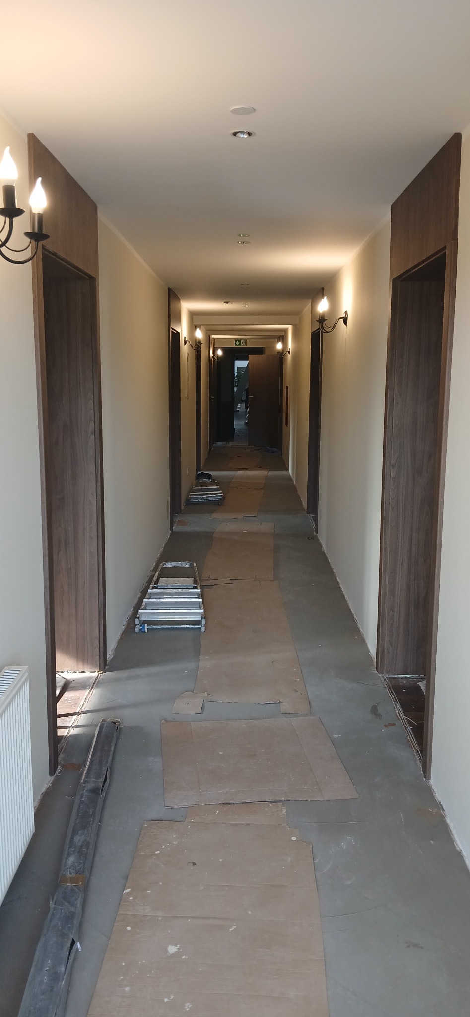 Korytarz w trakcie remontu: zabezpieczona podłoga kartonem, drabina, świeże ściany, nowe ościeżnice. Widok w głąb korytarza z lampami ściennymi.