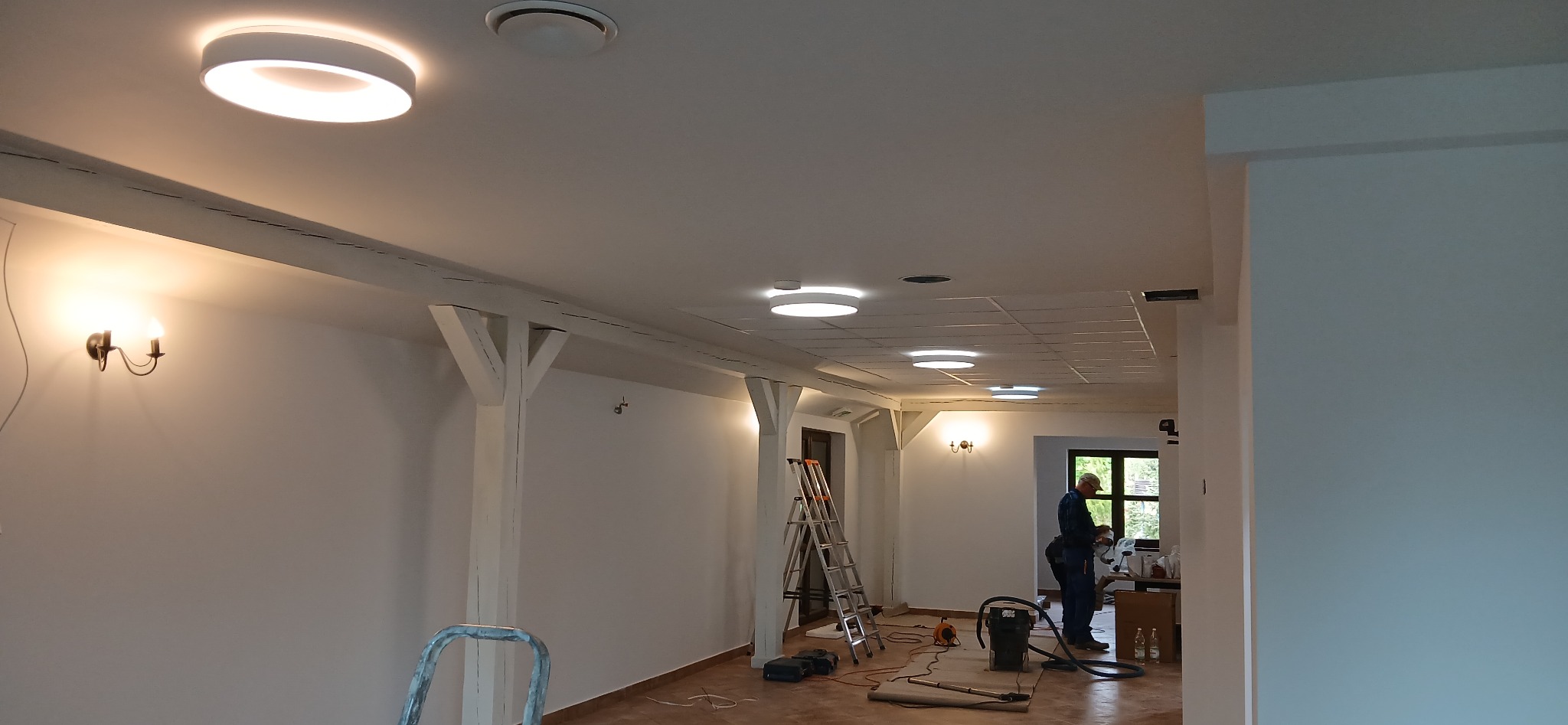 Wnętrze po remoncie: białe ściany z drewnianymi belkami, nowoczesne oświetlenie sufitowe, drabina i pracownik z odkurzaczem. Widoczne detale wykończenia.