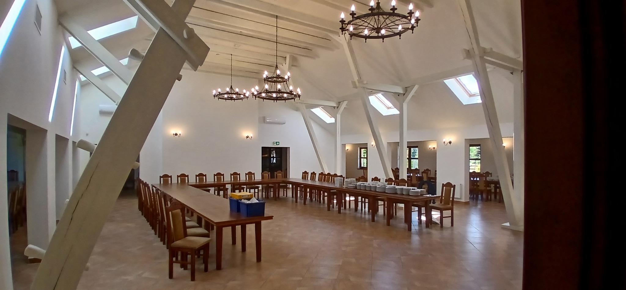 Przestronne wnętrze sali bankietowej z drewnianymi stołami w kształcie litery U, krzesłami i dekoracyjnymi żyrandolami. Białe ściany i strop z drewnianymi belkami.