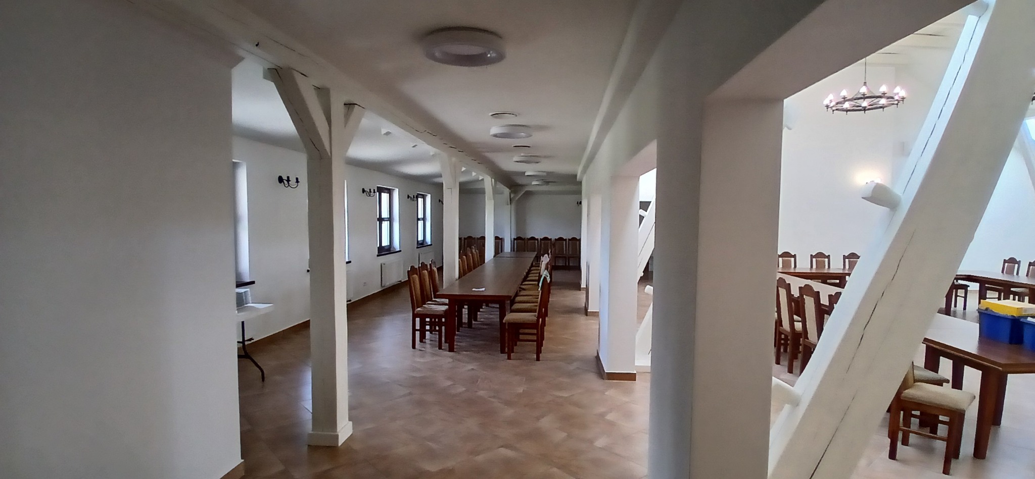 Jasne, przestronne wnętrze sali z drewnianymi stołami i krzesłami, po remoncie. Białe ściany i filary z drewnianymi elementami. Widoczne oświetlenie sufitowe i okna.