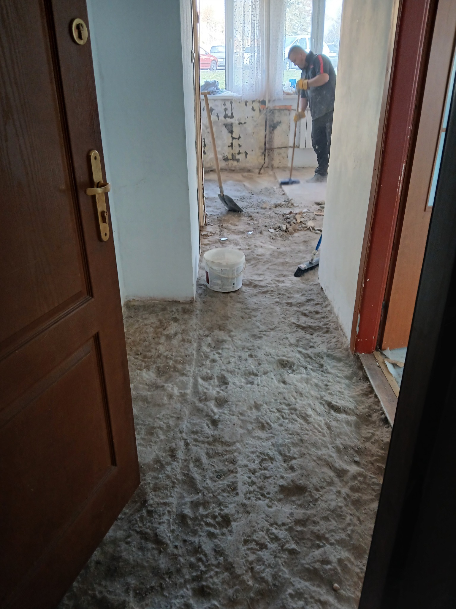 Remont mieszkania: widok przez otwarte drzwi na pomieszczenie w trakcie szpachlowania. Pracownik wyrównuje ścianę. Na podłodze warstwa pyłu i resztek materiałów budowlanych.