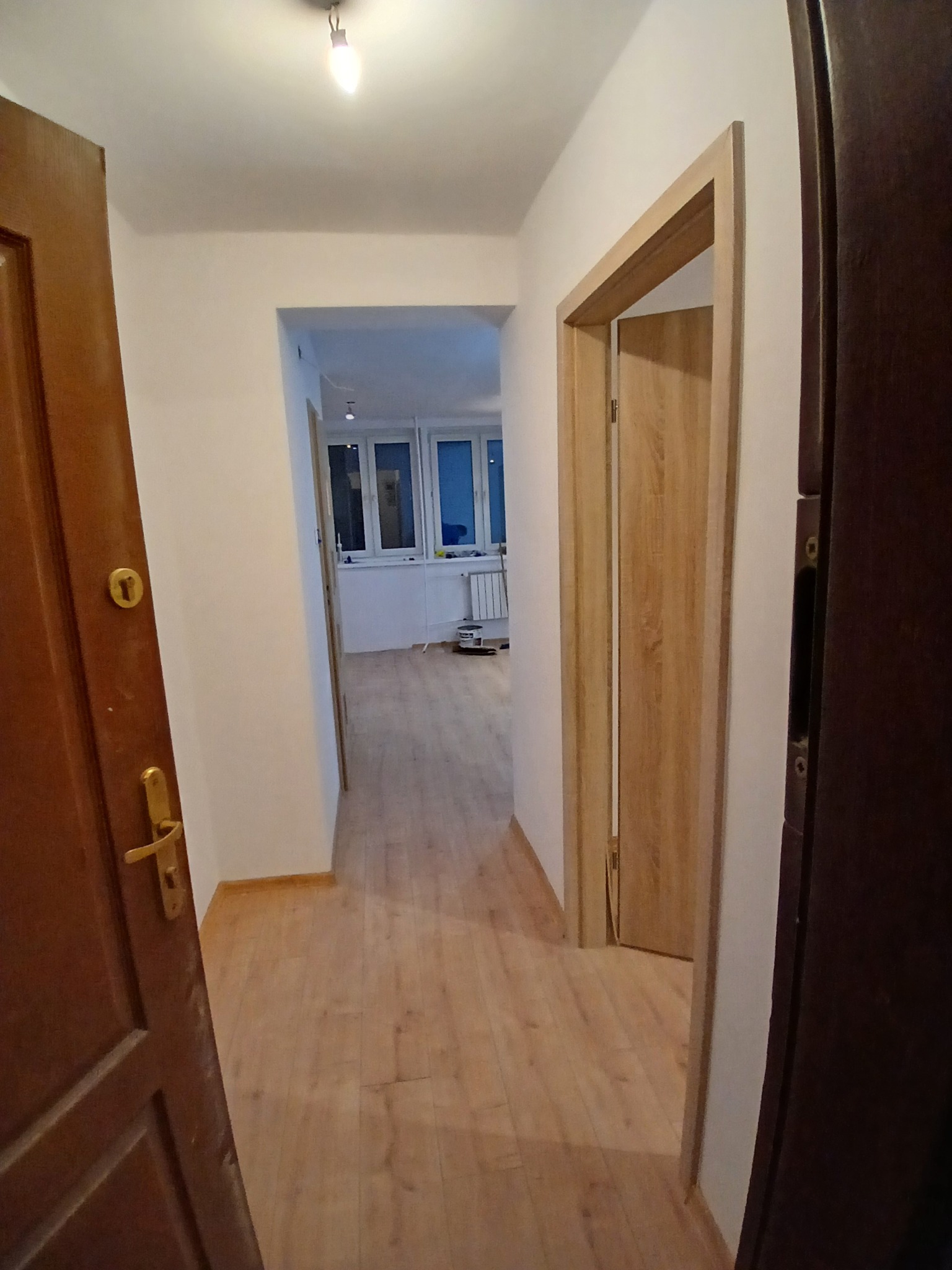 Jasne panele podłogowe w korytarzu, widoczne przez otwarte drzwi z drewnianą ościeżnicą. W głębi widoczny balkon z oknami.