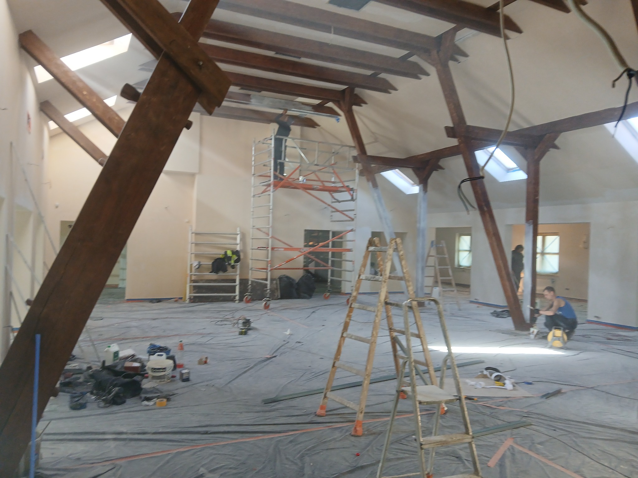 Wnętrze poddasza w trakcie remontu: widoczne rusztowania, drewniane belki stropowe, okna dachowe oraz narzędzia i materiały budowlane rozłożone na podłodze.