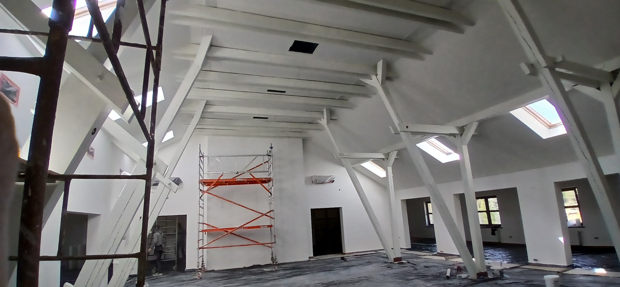 Wnętrze po malowaniu na biało, widoczne drewniane belki stropowe, rusztowanie malarskie i okna dachowe. Podłoga zabezpieczona folią.