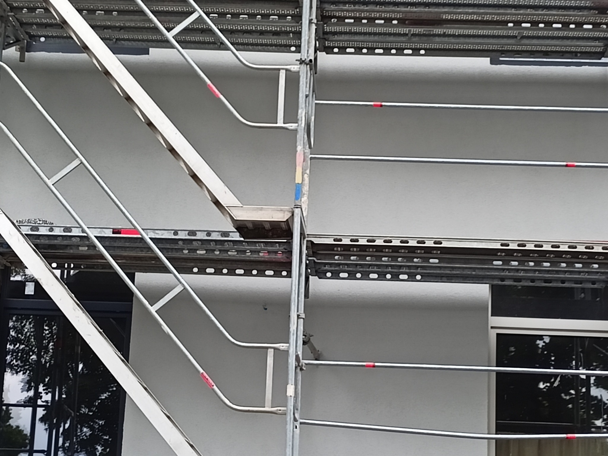 Elewacja budynku z oknami, częściowo zasłonięta metalowym rusztowaniem z czerwonymi oznaczeniami.