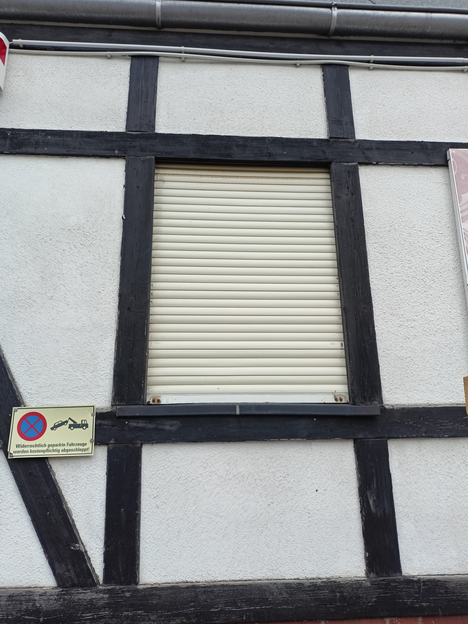 Fasada budynku w stylu szachulcowym z zamkniętą roletą zewnętrzną i znakiem zakazu parkowania.