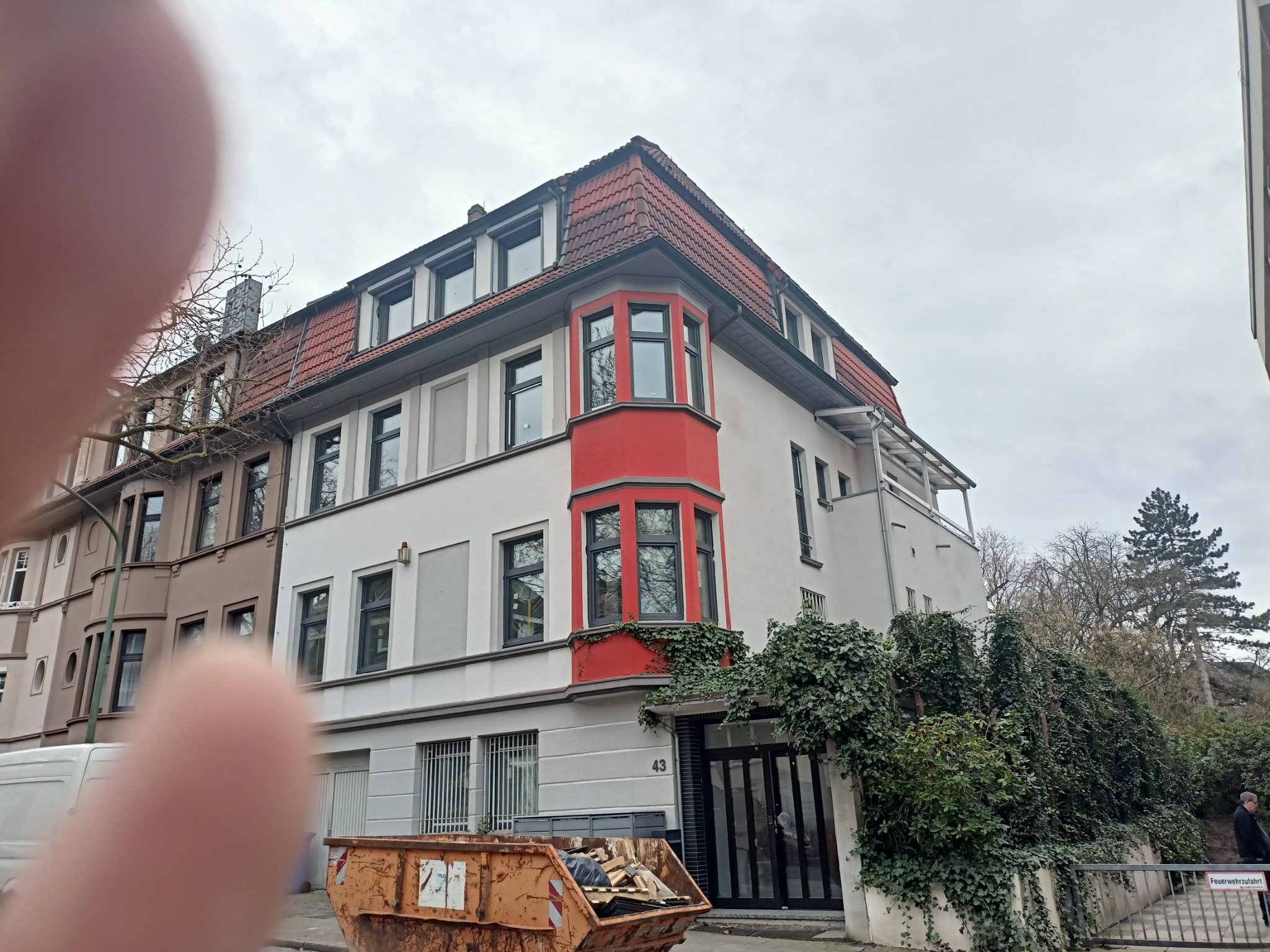 Budynek mieszkalny z czerwonym narożnym wykuszem okiennym, widok z dołu z fragmentem palca w kadrze, kontener na śmieci przed budynkiem.