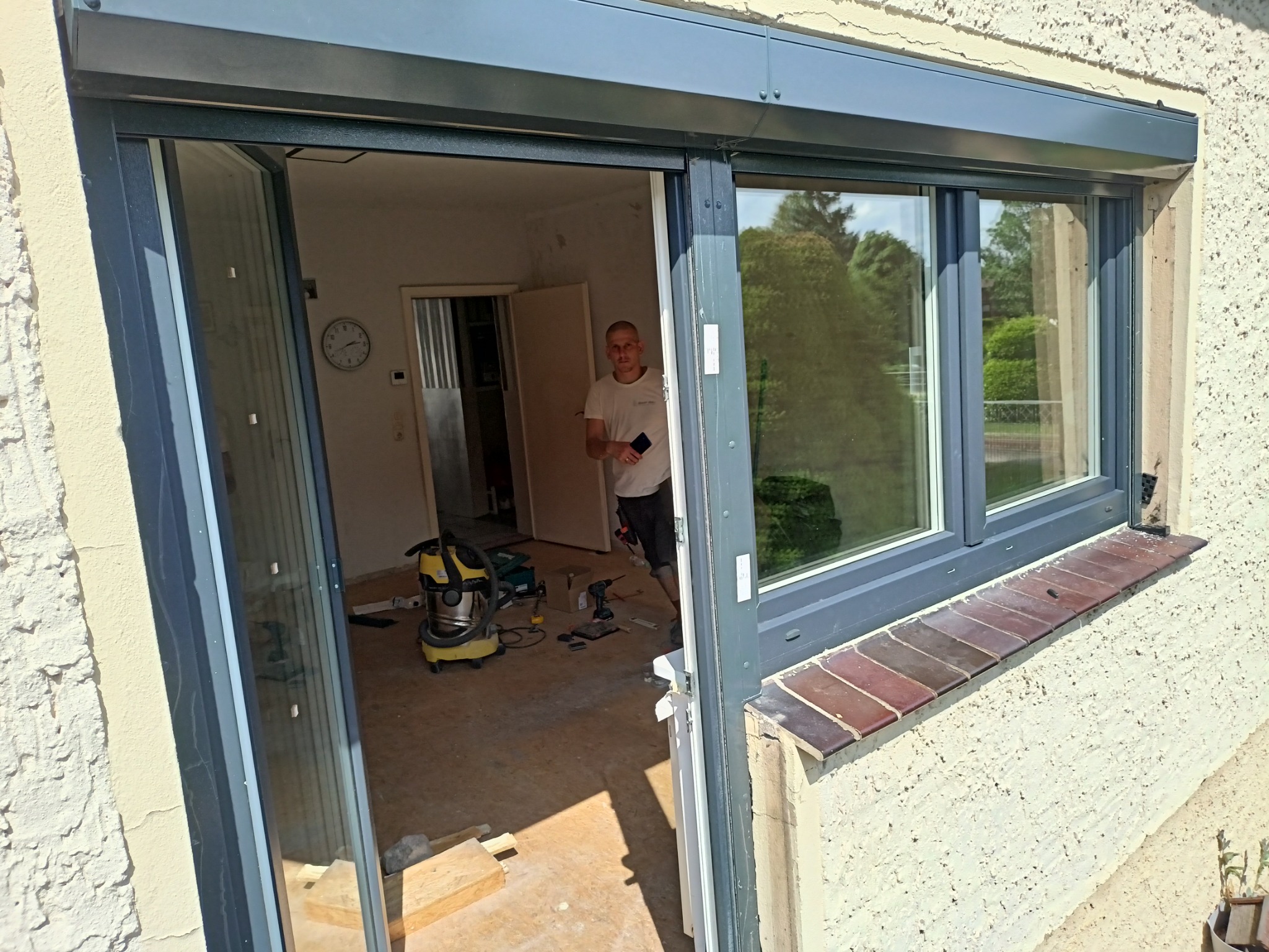 Montaż okna z roletą zewnętrzną: widok z zewnątrz na częściowo zamontowane okno z szarą ramą i zwiniętą roletą; w tle widoczny pracownik z telefonem oraz narzędzia.