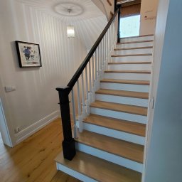 ZAKŁAD STOLARSKI KALETA PIOTR - Drewniane schody z białymi podstopnicami i czarną poręczą w jasnym wnętrzu. Widoczne drewniane podłogi i dekoracyjne oświetlenie sufitowe.