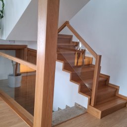 ZAKŁAD STOLARSKI KALETA PIOTR - Jasne, drewniane schody z przeszkloną balustradą i solidnym słupem nośnym, prowadzące na wyższą kondygnację. Widoczne detale wykończenia i naturalna kolorystyka drewna.