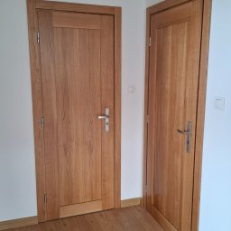 ZAKŁAD STOLARSKI KALETA PIOTR - Dwie drewniane, fornirowane drzwi wewnętrzne z prostymi ościeżnicami i srebrnymi klamkami, widoczne na tle białych ścian i podłogi z paneli w odcieniach dębu.