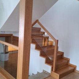 ZAKŁAD STOLARSKI KALETA PIOTR - Jasne, drewniane schody z przeszkloną balustradą i solidnym słupem nośnym, prowadzące na wyższą kondygnację. Widoczny fragment parkietu i schodów z innego materiału.
