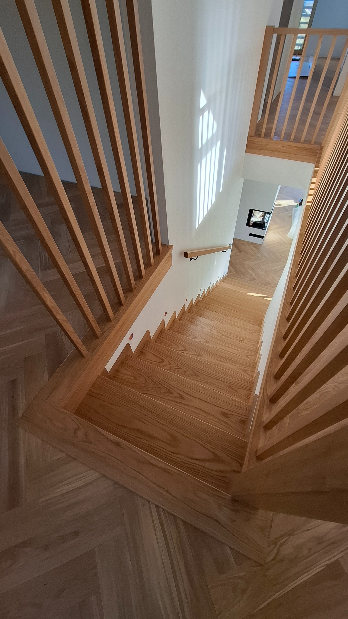 Jasne drewniane schody z barierką z pionowych listew, widok z góry, parkiet w jodełkę na podłodze, światło słoneczne na ścianie.