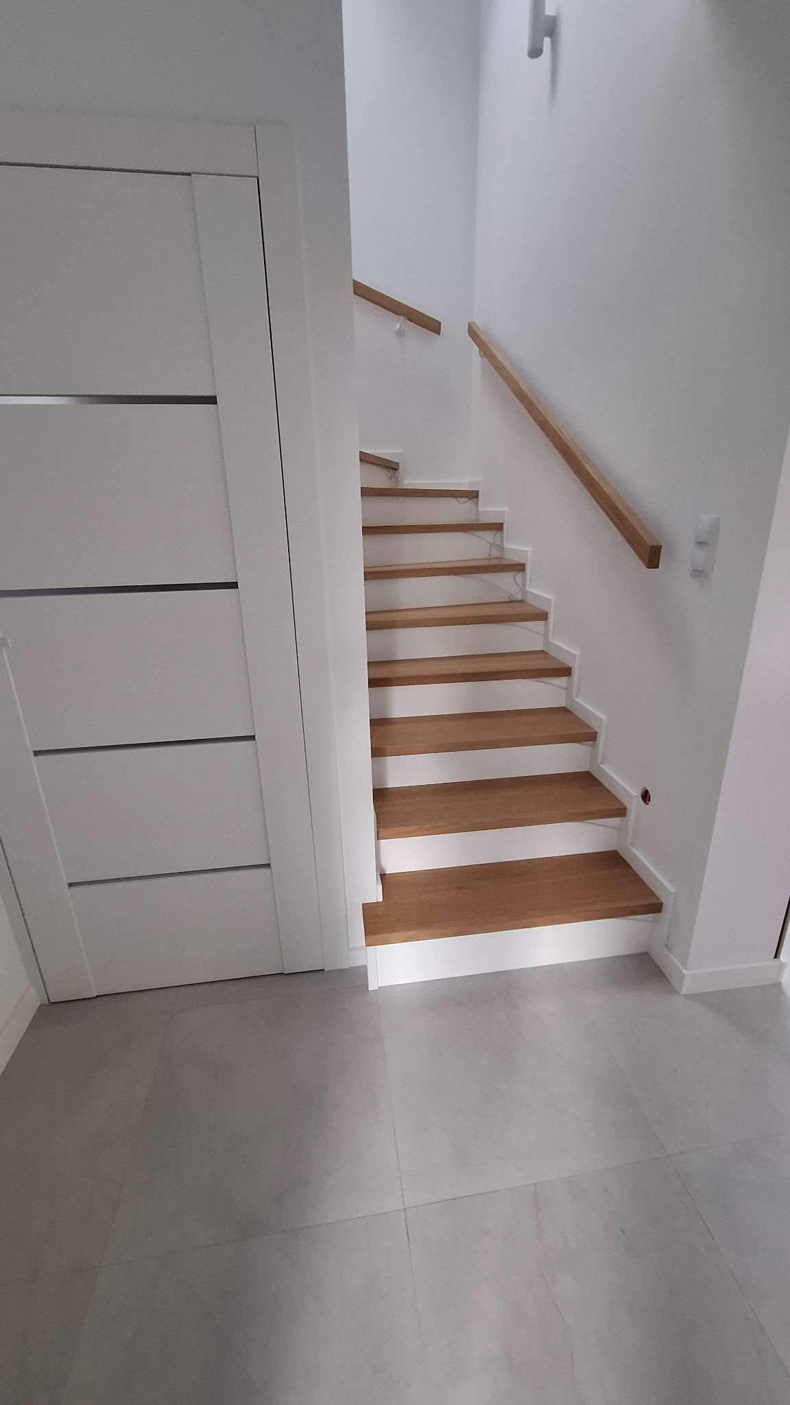 Jasne, minimalistyczne schody z drewnianymi stopniami i białą balustradą, oświetlone światłem z góry, widoczne z dołu.