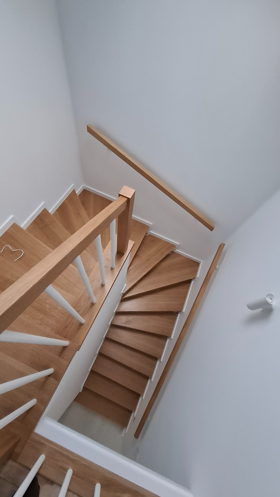 Widok z góry na jasne, drewniane schody z białymi balustradami w minimalistycznym wnętrzu z białymi ścianami i oświetleniem punktowym.