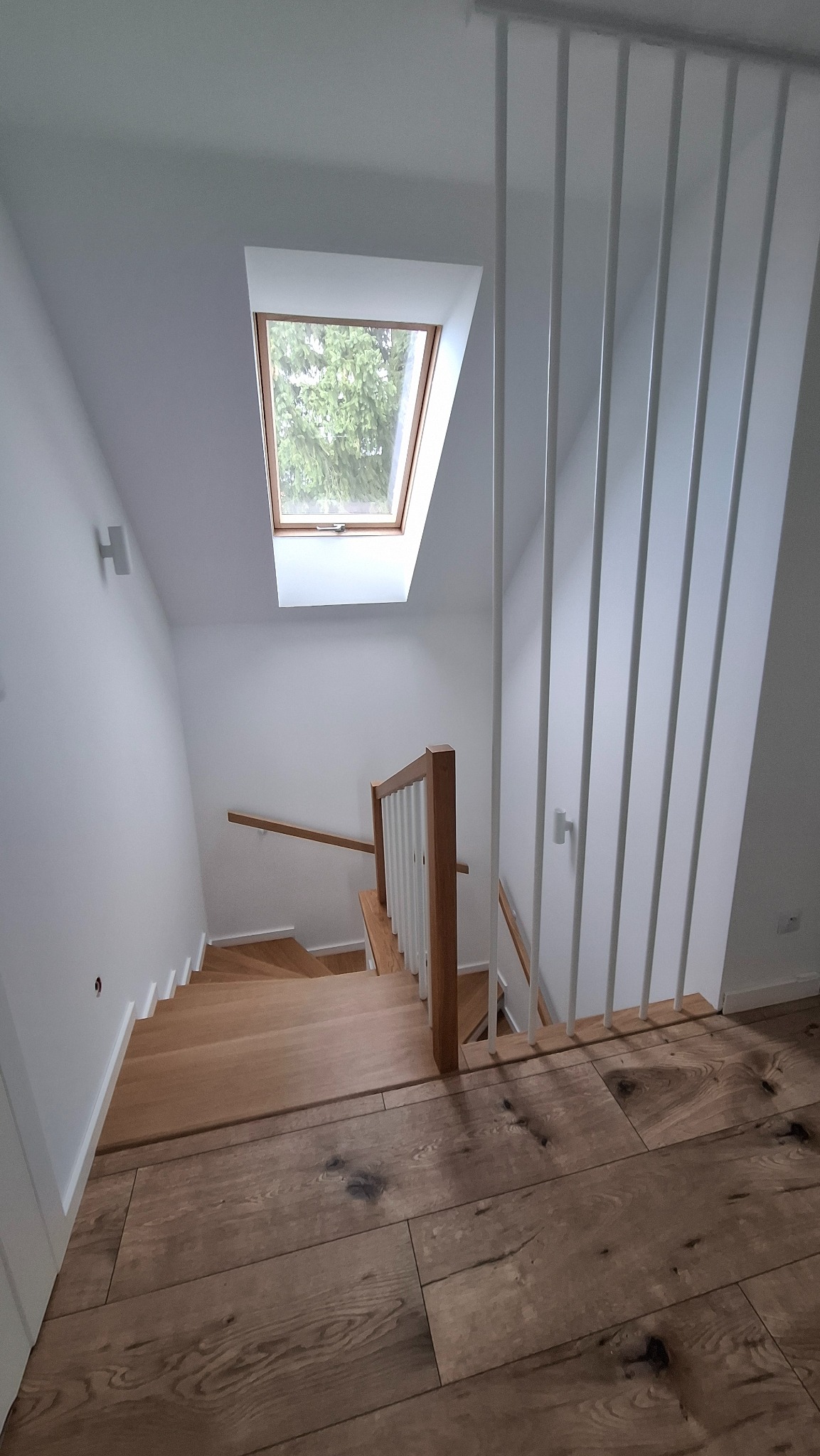 Widok z góry na nowoczesne, drewniane schody z białymi, pionowymi balustradami w domu w Skrzydlnej, oświetlone naturalnym światłem z okna dachowego.