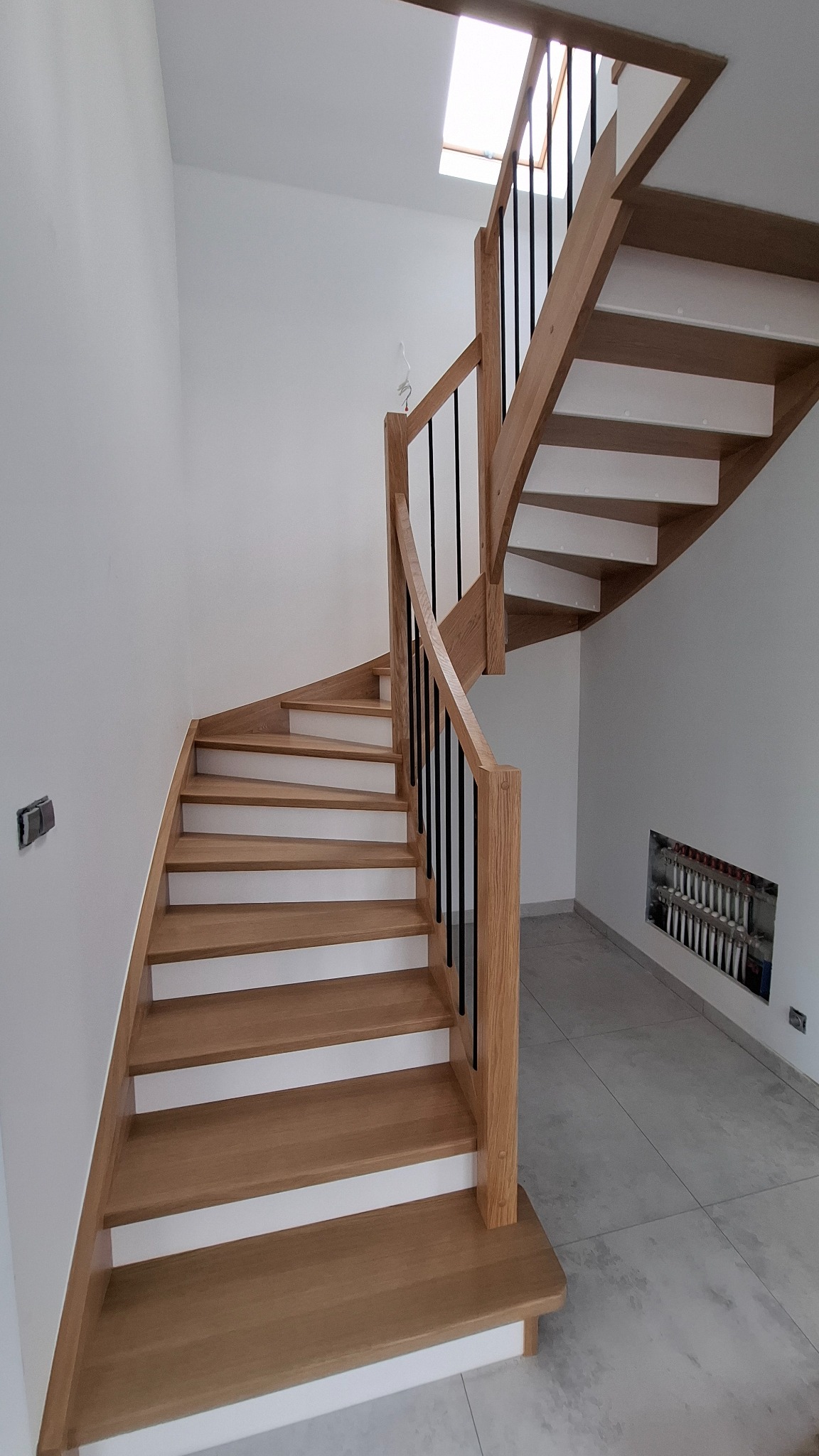 Drewniane schody gięte z białymi podstopnicami i czarnymi balustradami w nowoczesnym wnętrzu z oknem dachowym.