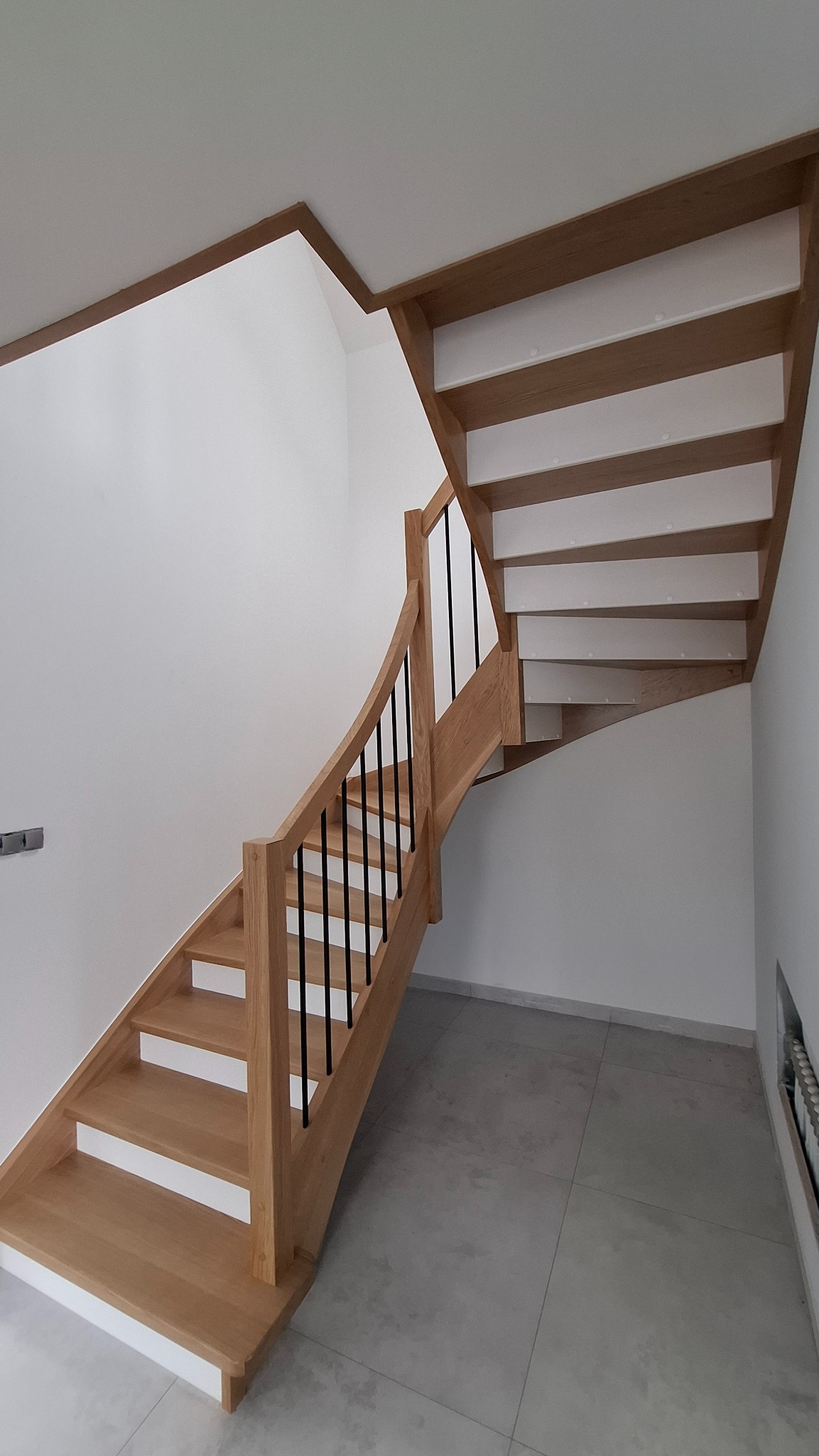 Drewniane schody z giętą poręczą i czarnymi tralkami, białe podstopnice, jasne drewno, szara podłoga z płytek.
