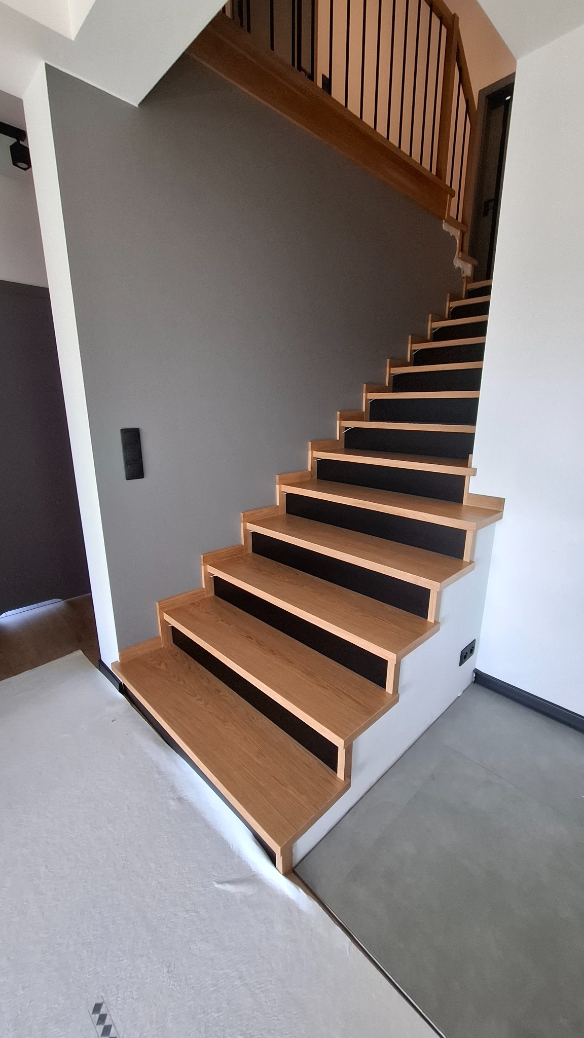 Nowoczesne schody z drewnianymi stopniami i czarnymi podstopnicami, widok z dołu na szarą ścianę i białe wykończenia.