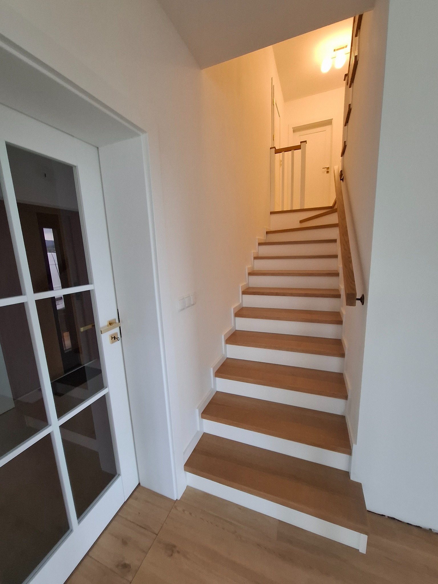 Jasne drewniane schody z białymi podstopnicami prowadzące na górę, widoczne z poziomu podłogi z drewnianych paneli, obok białe drzwi z szybami i złotą klamką.