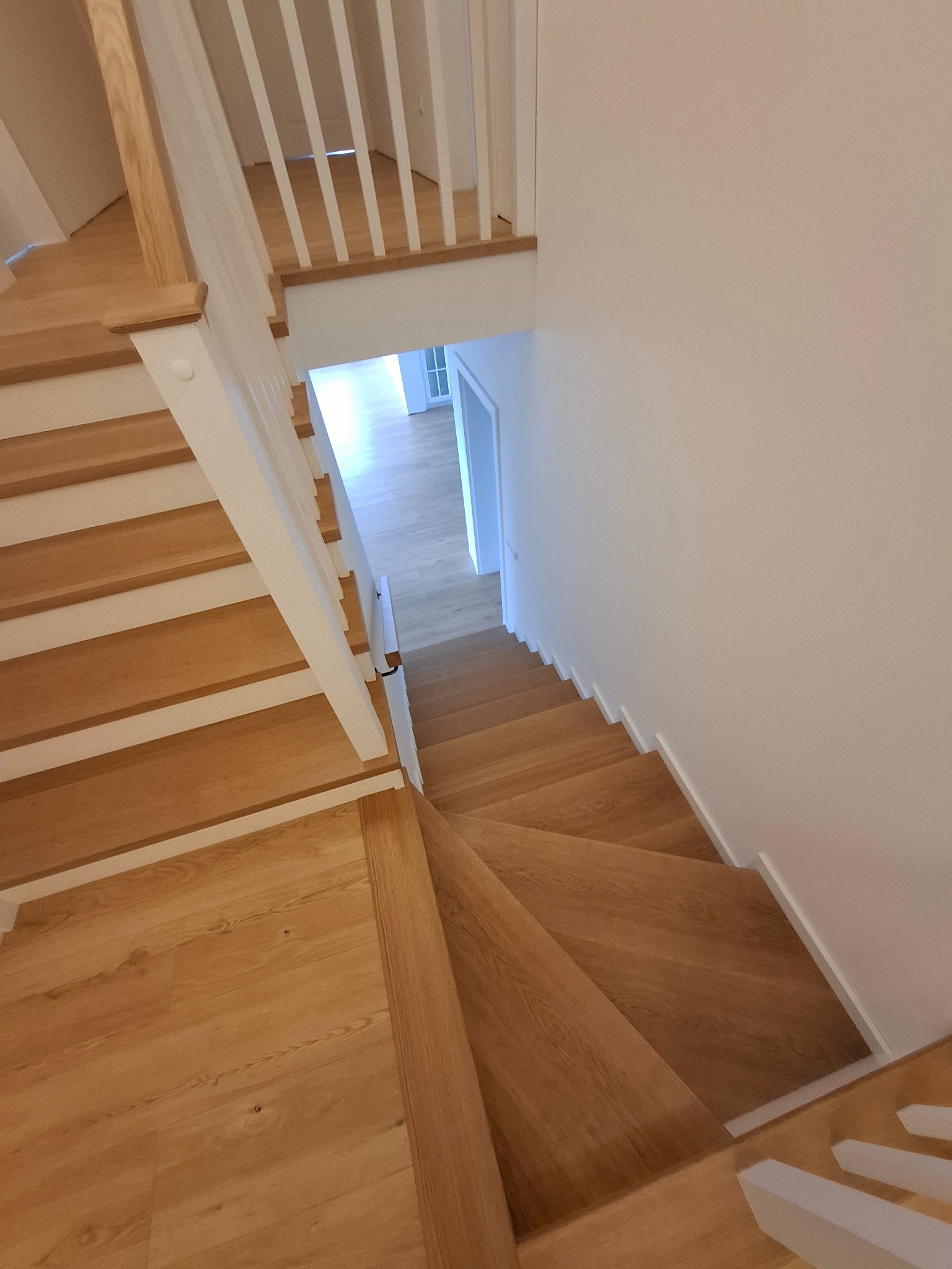 Widok z góry na jasne, drewniane schody z białymi podstopnicami i balustradą, prowadzące w dół do jasnego pomieszczenia.