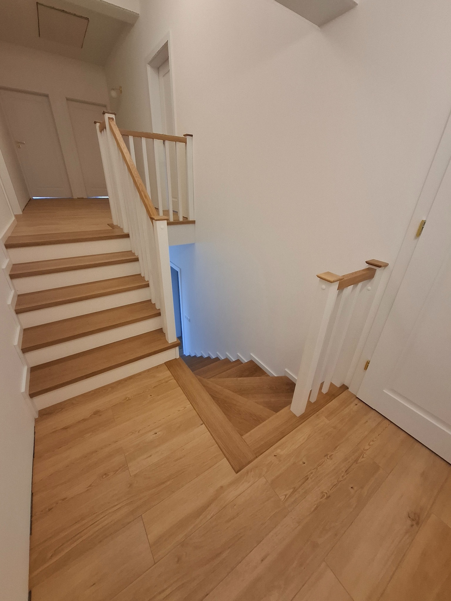 Jasne, drewniane stopnie z białymi podstopnicami i balustradą, prowadzące w dół, widok z góry, naturalne oświetlenie.