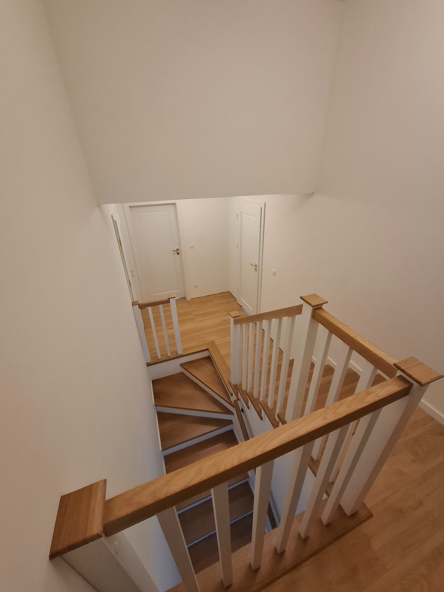 Widok z góry na drewniane schody z białymi balustradami w jasnym wnętrzu, dwie białe drzwi widoczne w tle.