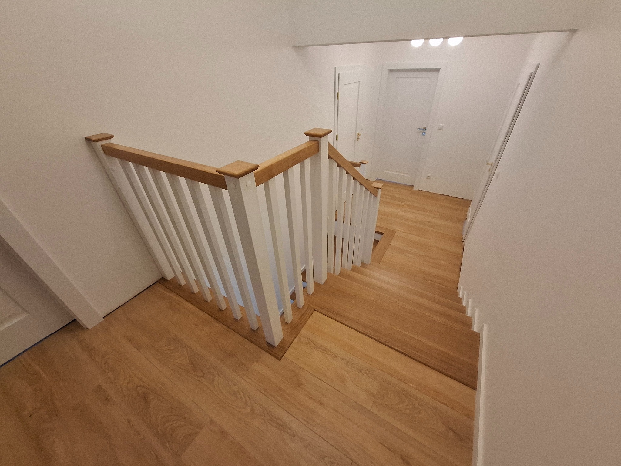 Jasne drewniane schody z białymi balustradami i dębową poręczą w nowoczesnym wnętrzu z białymi ścianami i drzwiami.