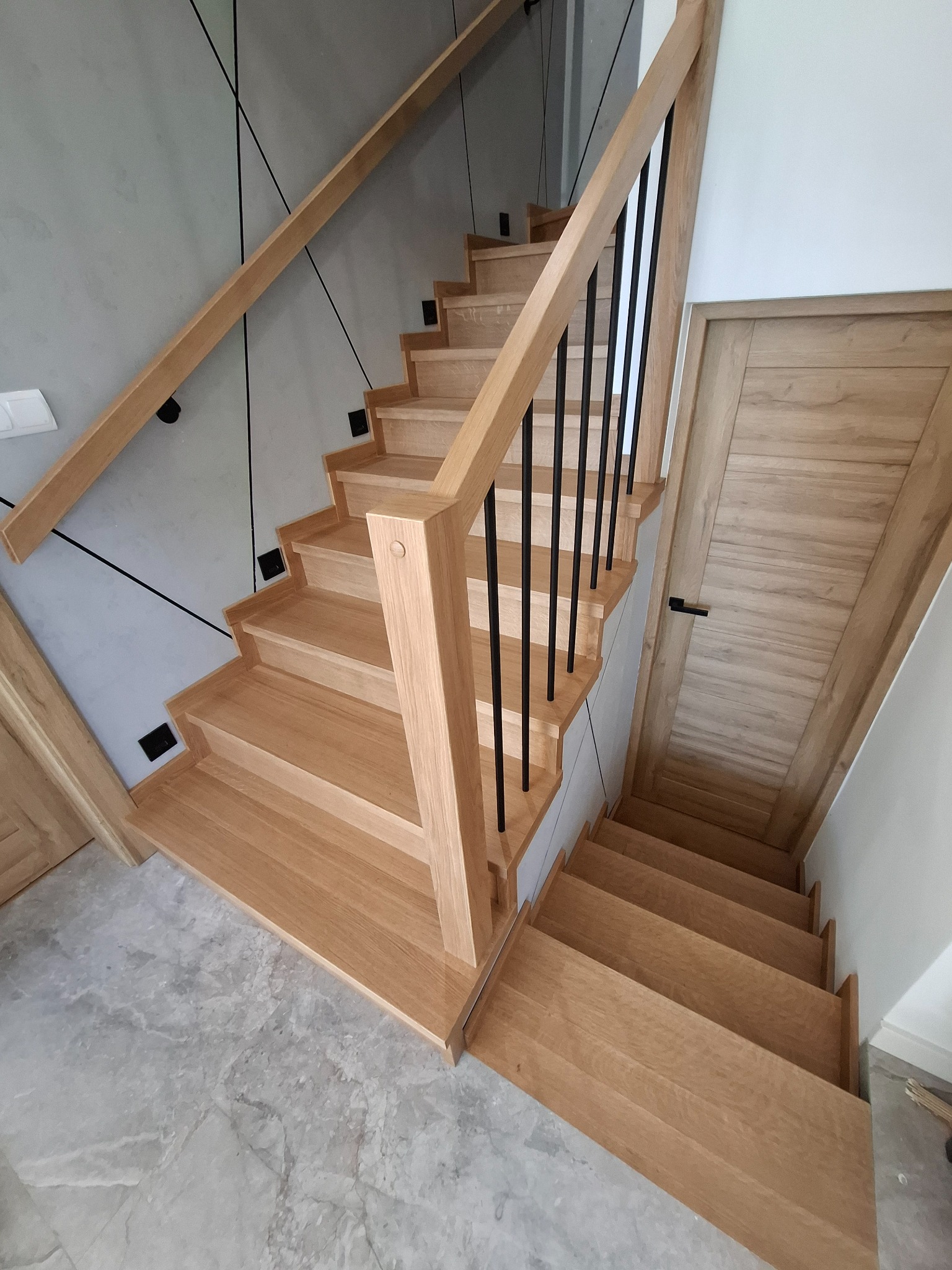 Jasne, dębowe stopnie z czarnymi, metalowymi prętami balustrady i dopasowaną kolorystycznie boazerią ścienną, widok z góry na dół.