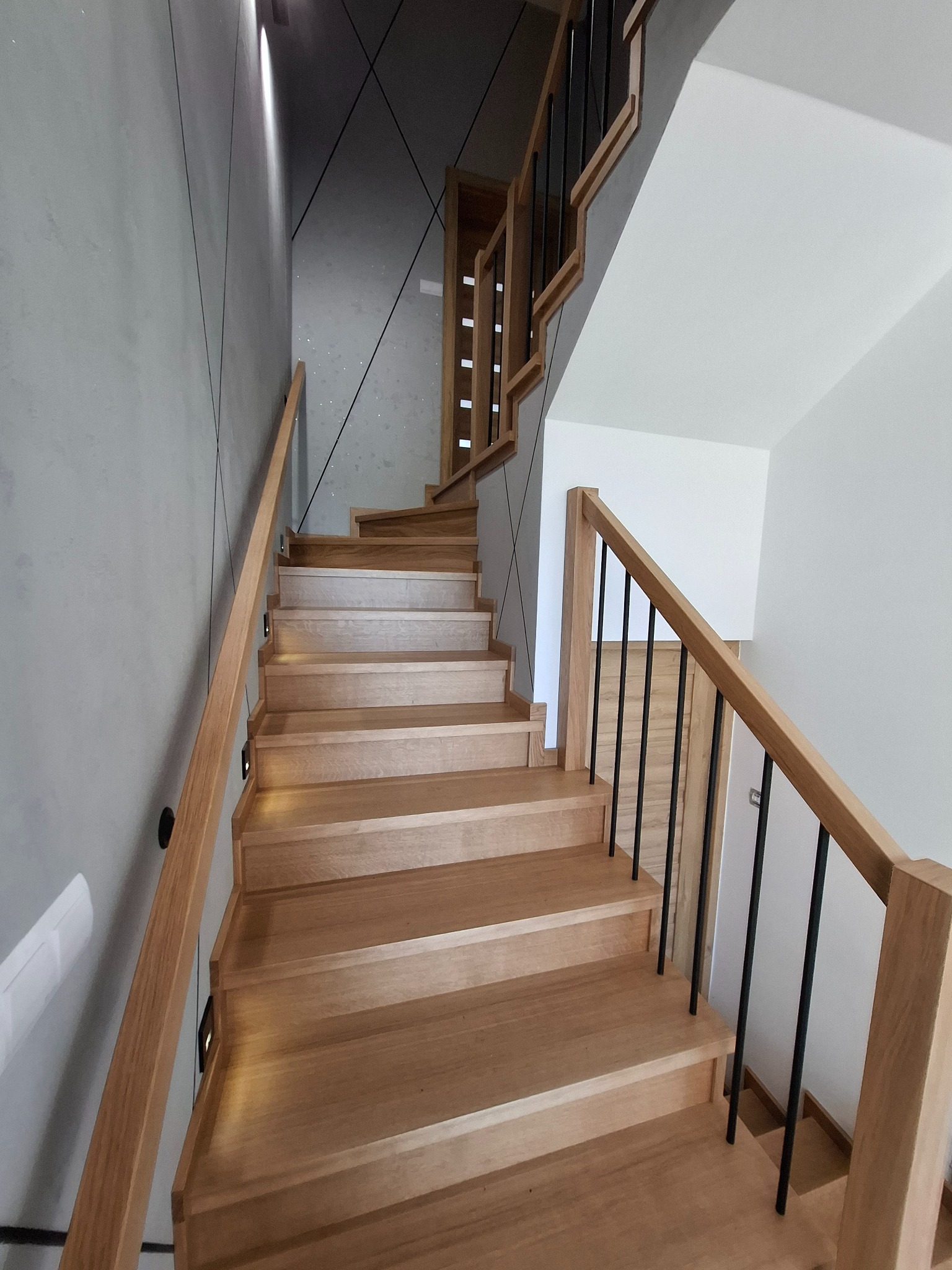 Jasne, drewniane stopnie z podświetleniem i czarnymi, metalowymi prętami balustrady, umieszczone w nowoczesnym wnętrzu z szarą ścianą o geometrycznym wzorze.