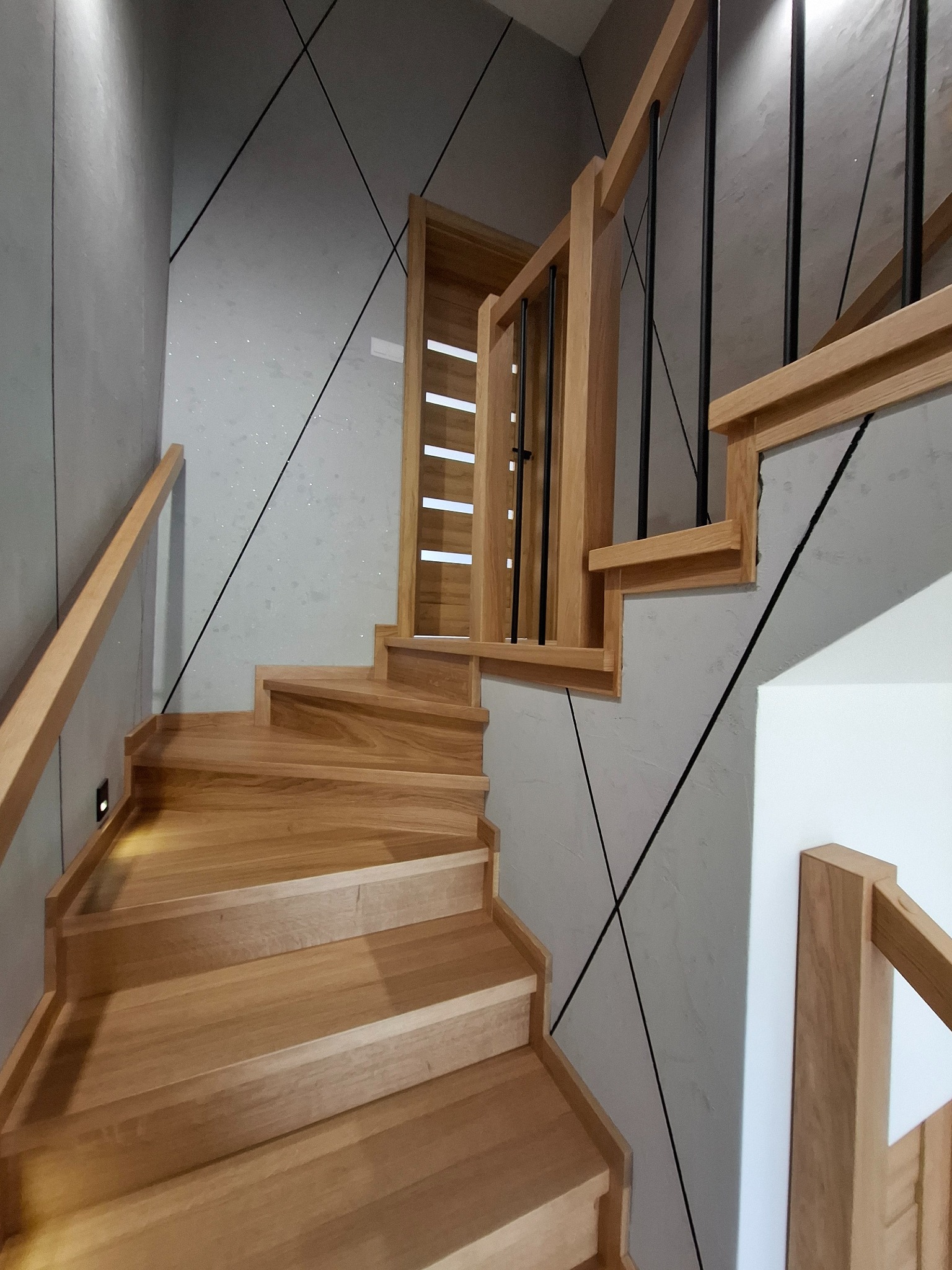 Drewniane schody z poręczą z czarnymi, metalowymi prętami, oświetlenie LED przy stopniach, ściana z geometrycznym wzorem.