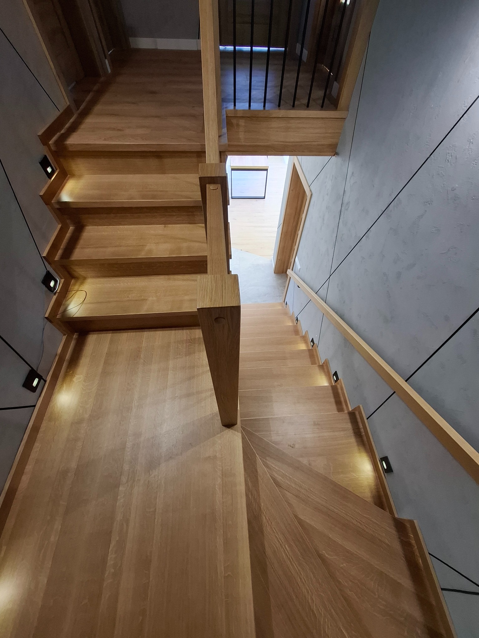 Wewnętrzne, dębowe schody z minimalistyczną balustradą, oświetleniem punktowym w stopniach i szarą ścianą z geometrycznym wzorem.